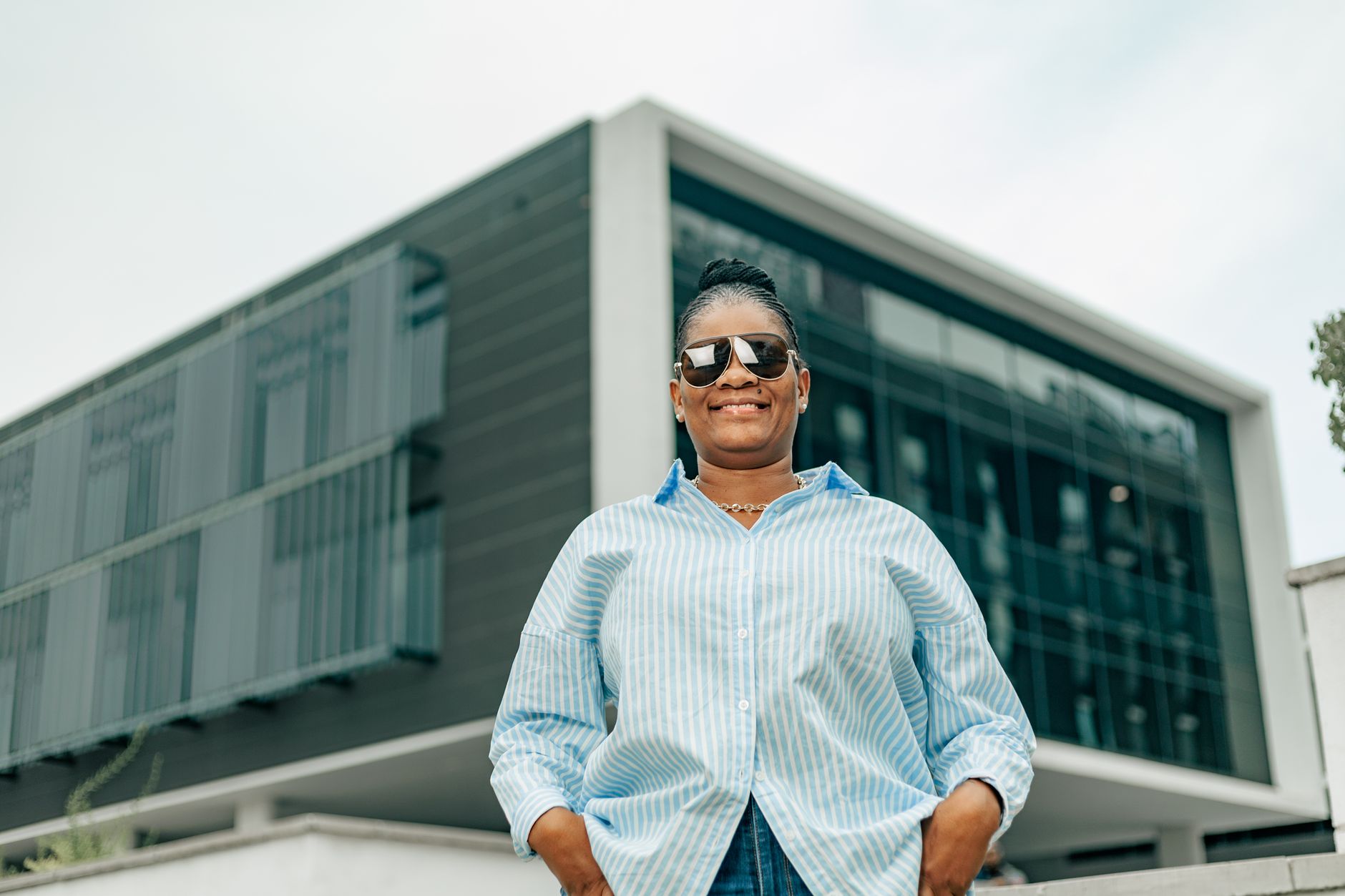 Woman in striped shirt posing confidently in front of modern architecture on a sunny day.
