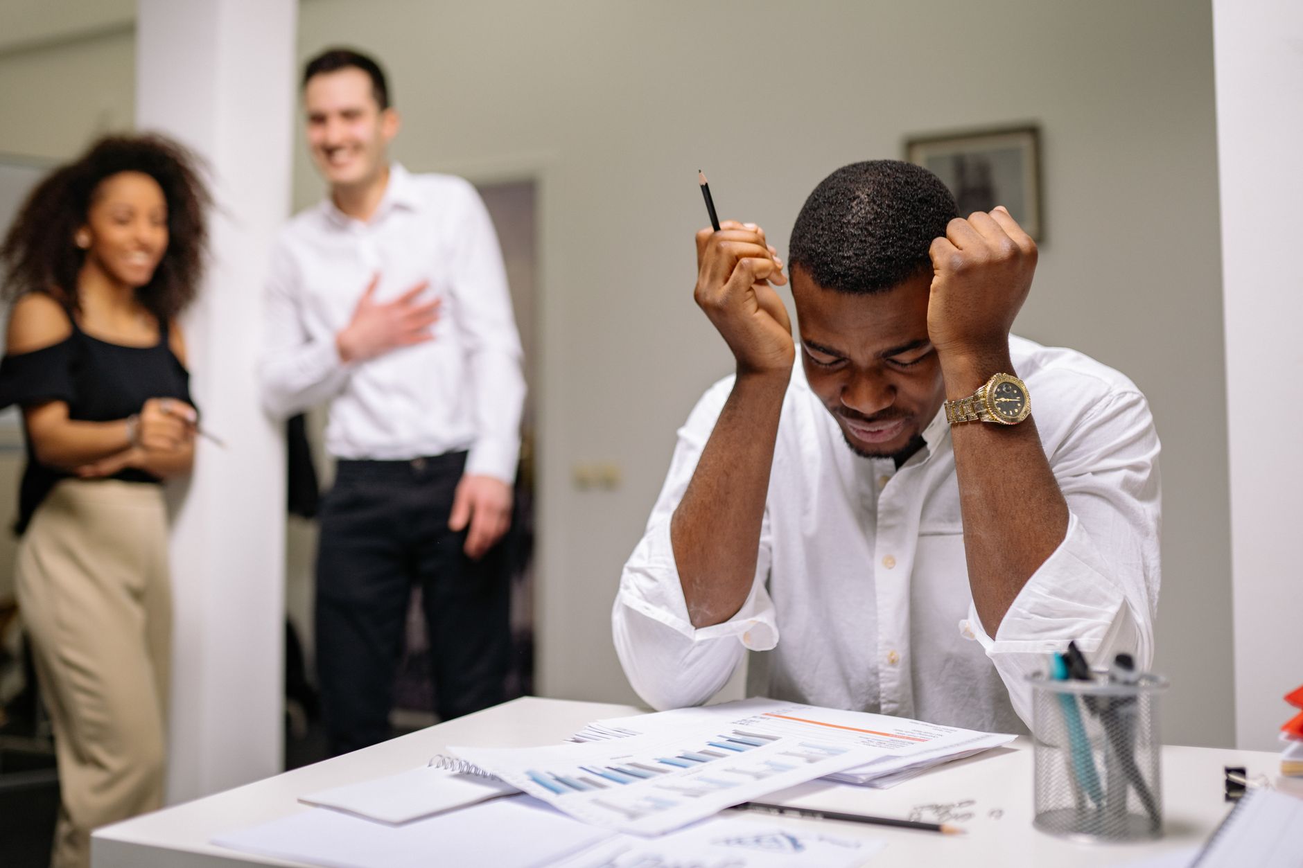 Stressed man at an office desk surrounded by colleagues, feeling overwhelmed at work.