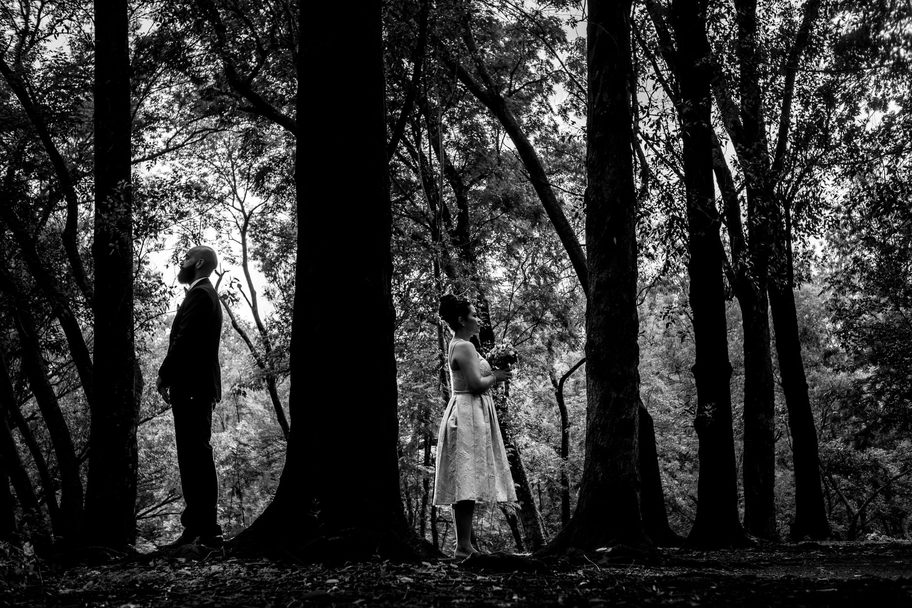 Dramatic black and white portrait of a couple standing back to back in a forest setting.