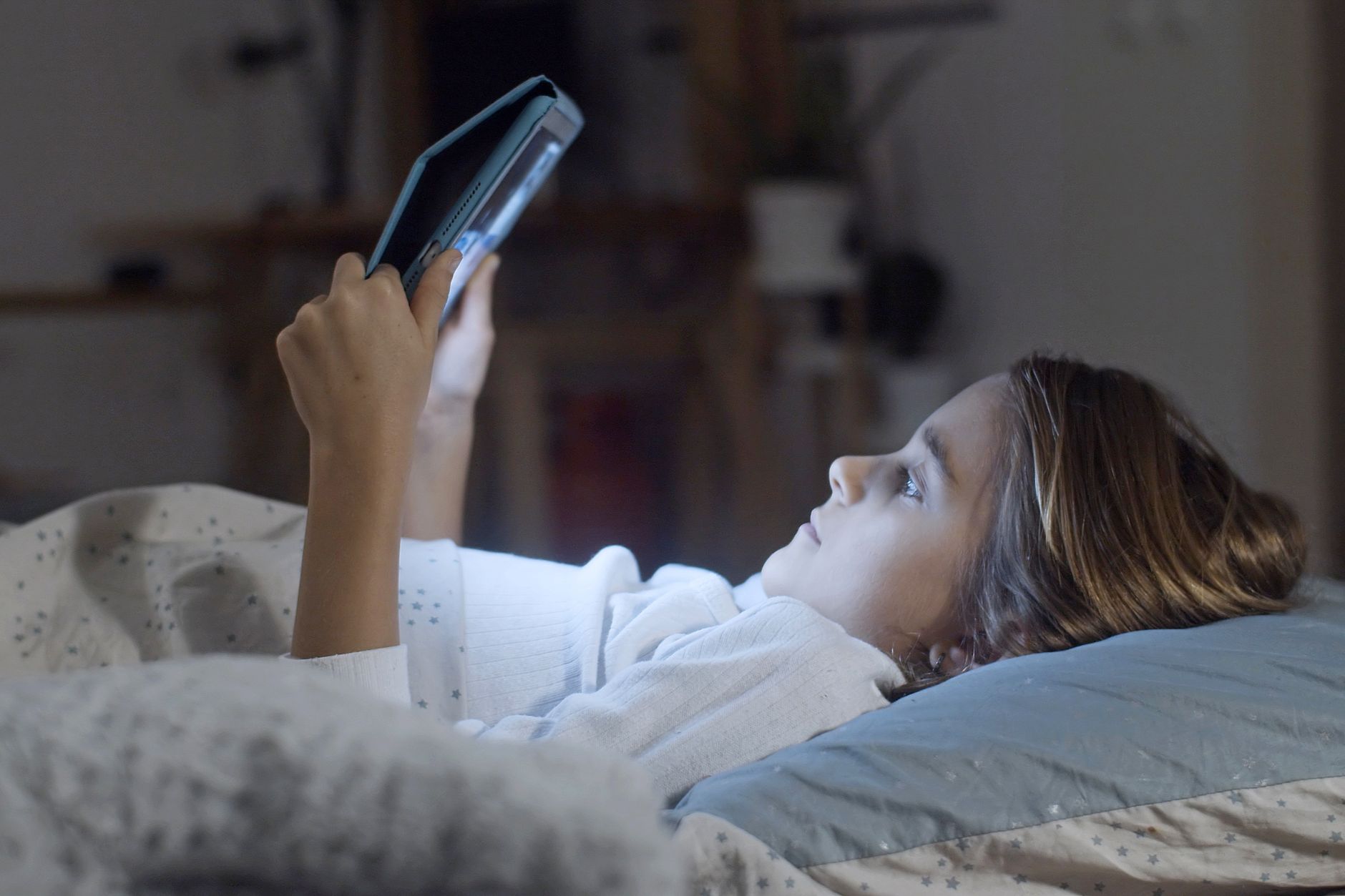 A young girl lying in bed using a tablet during nighttime indoors.