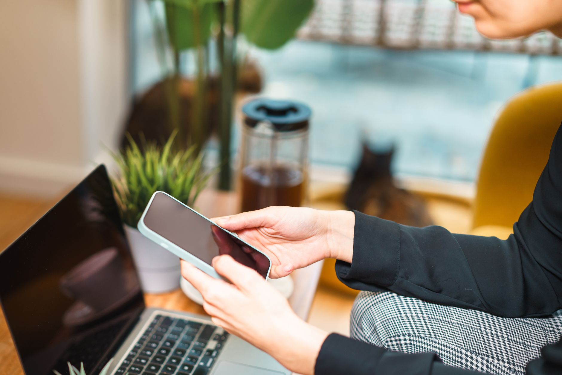 Woman working remotely from home, using a smartphone and laptop at a cozy desk with a plant and cat in background.