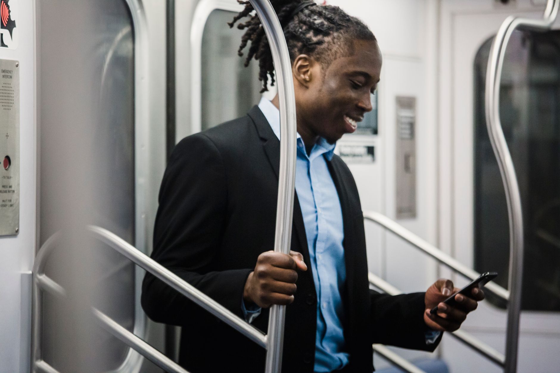 Young African American male in elegant office suit browsing social media on phone and smiling while going home by subway train