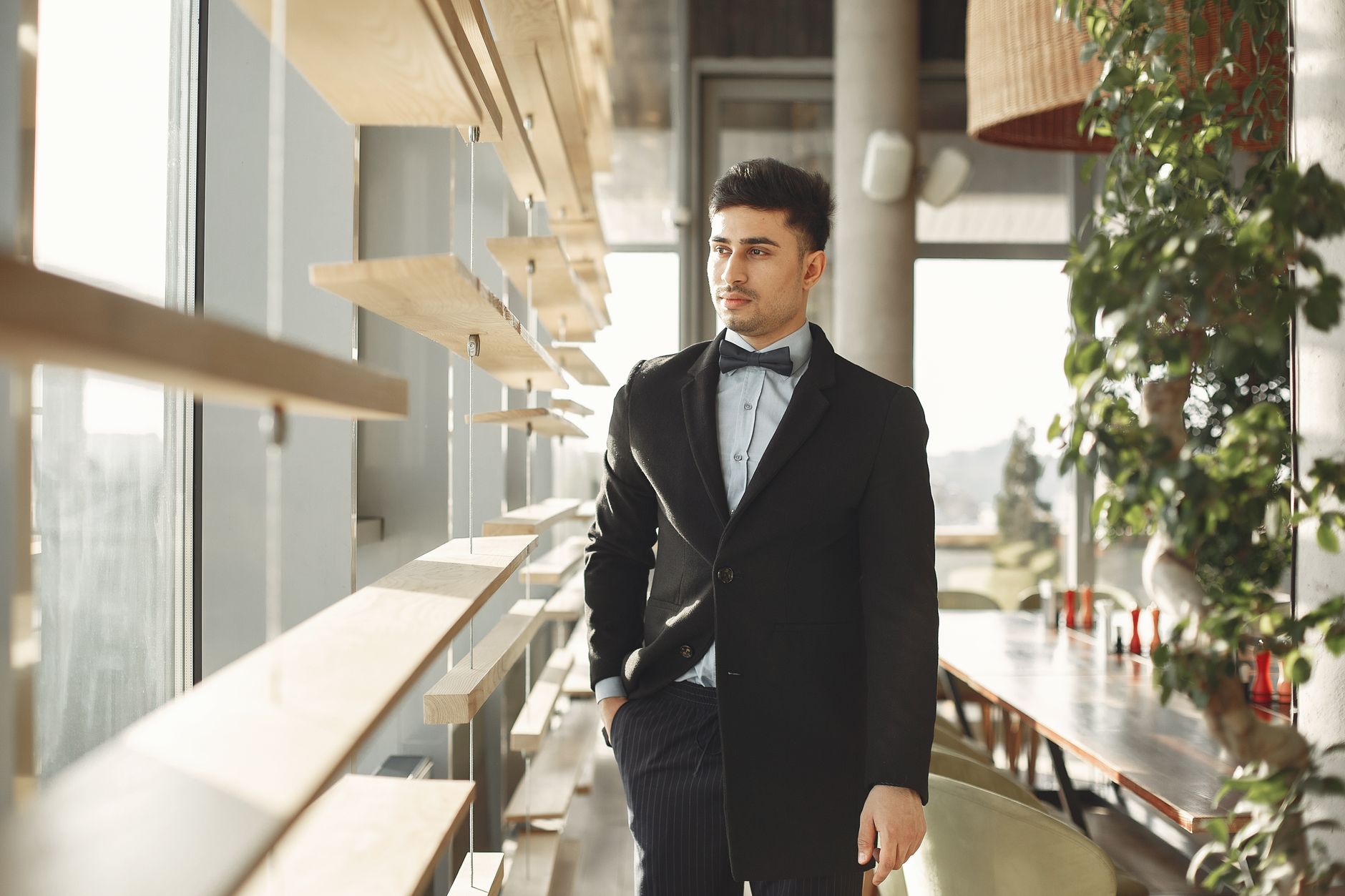 Stylish man in a formal suit with a bow tie standing indoors by a sunlit window.